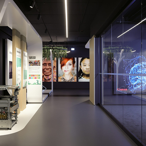 Ein moderner Ausstellungsraum mit Informationsdisplays, großen Porträts von verschiedenen Gesichtern an der Rückwand und einer leuchtenden digitalen Gehirngrafik hinter Glas auf der rechten Seite. Der Bereich ist hell beleuchtet und klar und minimalistisch gestaltet.