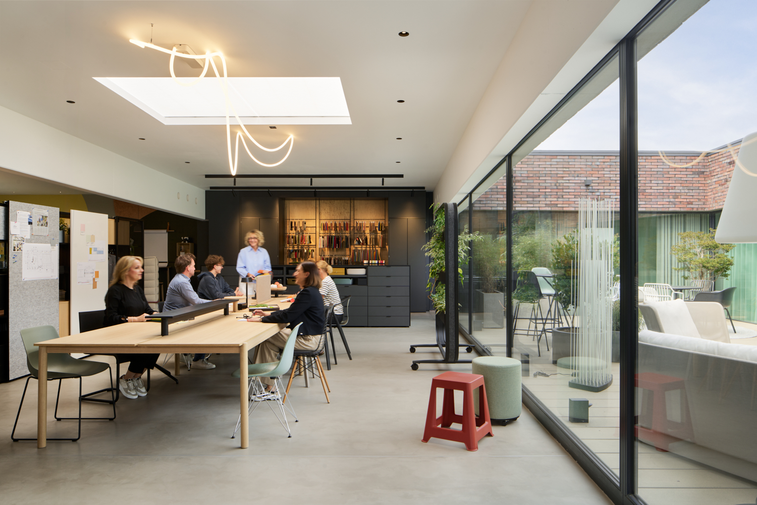 Eine Gruppe von Menschen sitzt um einen Holztisch herum in einem modernen, hellen Büroraum mit großen Glasfenstern, einem Oberlicht und minimalistischen Möbeln. Im Hintergrund stehen zwei Personen in der Nähe eines Küchenbereichs.