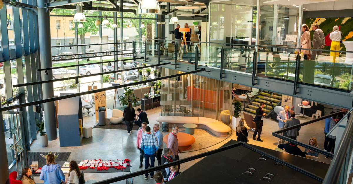 Ein modernes Großraumbüro mit zwei Etagen, in dem Menschen zusammenkommen, arbeiten und spazieren gehen. Es gibt Glaswände, Pflanzen, bunte Möbel und große Fenster, die natürliches Licht hereinlassen.