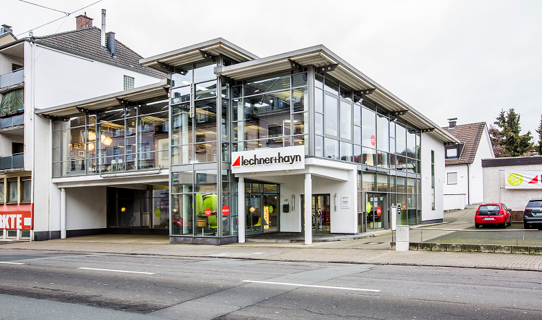 Ein modernes Gebäude mit Glasfront und Lechnerhoyn-Schriftzug; mehrere Autos sind seitlich geparkt, und im Vordergrund sind die Straße und der Bürgersteig zu sehen.
