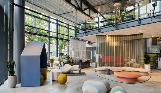 Moderne Büro-Lounge mit großen Fenstern, stilvollen Sitzgelegenheiten, Topfpflanzen und einem verglasten Zwischengeschoss. Geschwungene Sofas, Poufs und einzigartige Möbelstücke schaffen eine helle, luftige und einladende Atmosphäre.