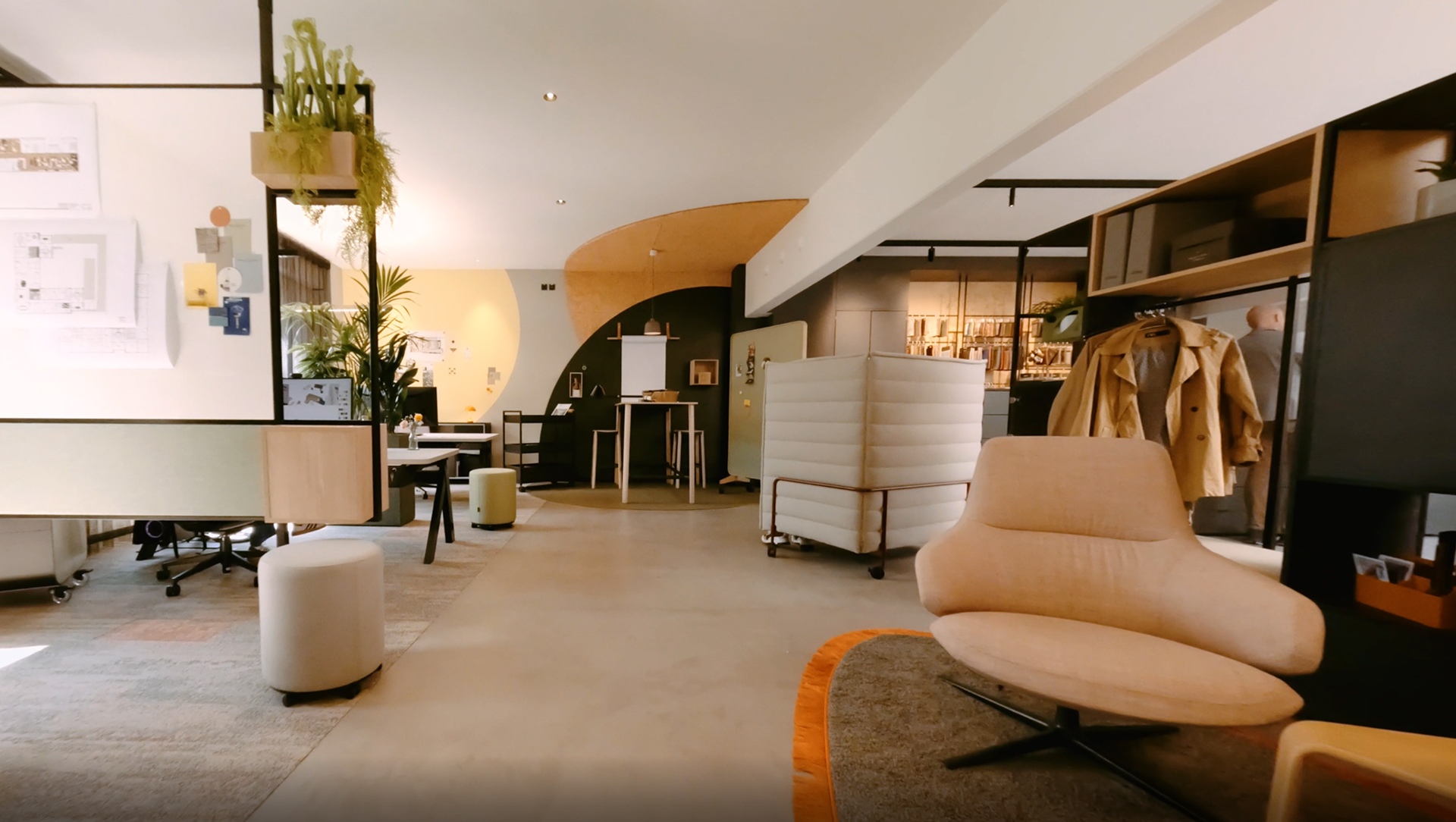 Ein modernes Großraumbüro mit einem beigen Loungesessel, einem abgetrennten Arbeitsbereich, hängenden Pflanzen und einem Dekor in neutralen Farbtönen. Regale mit Mänteln, ein Whiteboard und verschiedene Sitzbereiche schaffen eine gemütliche und organisierte Atmosphäre.