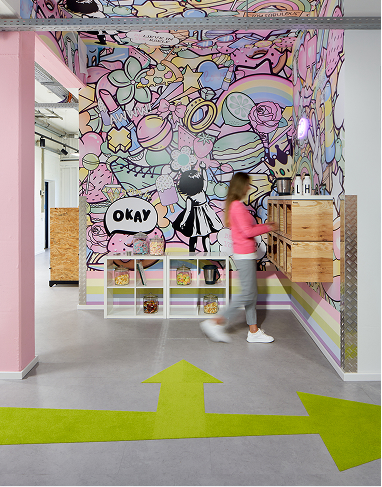 Eine Person geht an bunten Wandbildern im Cartoon-Stil vorbei, die Süßigkeiten, Sterne und verspielte Formen in einem modernen Raum mit Bonbongläsern in Regalen und leuchtend grünen Pfeilen auf dem grauen Boden zeigen.