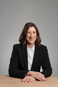 Eine Frau mit schulterlangen braunen Haaren, die einen schwarzen Blazer und ein weißes Hemd trägt, sitzt an einem Holztisch und lächelt in die Kamera vor einem schlichten grauen Hintergrund.